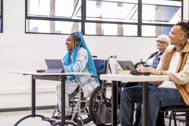 Photo: Students with laptops in a classroom setting, including a cheerful woman using a wheelchair and another student with short hair and glasses, in a bright room with large windows.